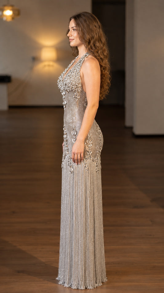 Side profile view of model posing in a silver rhinestone halter maxi dress with fringe skirt